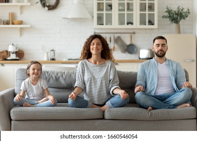Ruhige junge Familie mit kleiner Tochter sitzt auf der Couch und praktiziert gemeinsam Yoga, glückliche Eltern mit kleinen Vorschulkindern ruhen sich auf dem Sofa aus und meditieren, um negative Emotionen am Wochenende zu Hause zu lindern