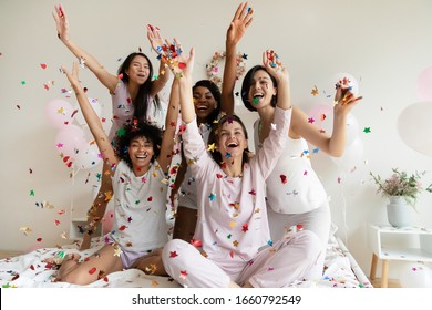 Entusiasmadas chicas diversas usando pijamas arrojando confeti colorido, gritando de alegría, celebrando en una fiesta de pijamas o en una despedida de soltera, cinco hermosas mujeres jóvenes divirtiéndose juntas en la cama en el dormitorio