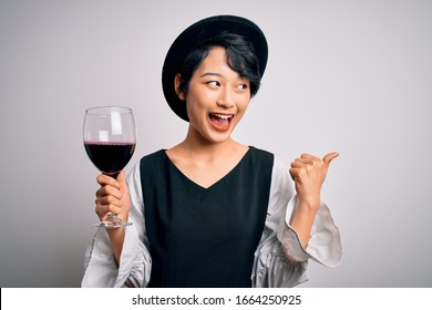 Joven y hermosa sommelier asiática bebiendo un vaso de vino tinto sobre un fondo blanco aislado señalando y mostrando con el pulgar hacia un lado con la cara feliz sonriendo