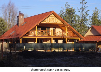 facade of a brown private house with a wooden balcony under a red tiled roof behind a green fence against a blue sky on a sunny day