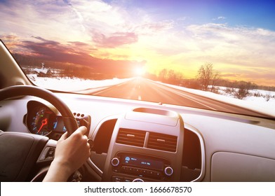 Autodashboard met de hand van de bestuurder op het zwarte stuur tegen een winterse asfaltweg in beweging en een nachtelijke hemel met een zonsondergang