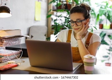 Jovem jardineira de óculos, usando laptop, se comunica pela internet com um cliente em seu jardim/estufa residencial, com uma caneca reutilizável de café/chá sobre a mesa. Ambiente de trabalho aconchegante, trabalho remoto, conceito de e-learning.