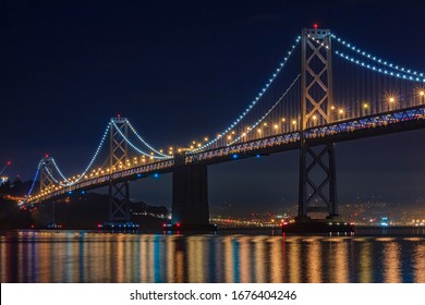 San Francisco Bay Bridge on a clear night, lit up by yellow and blue lights, reflecting of the water in the Bay, long exposure