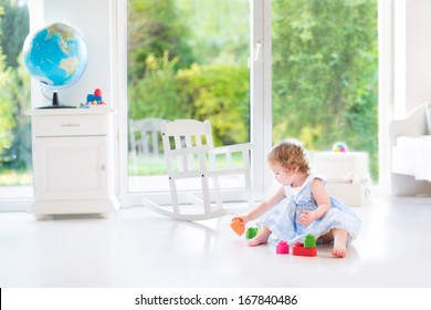 庭の景色を望む大きな窓のある白い日当たりの良い寝室で遊ぶ青いドレスを着た巻き毛のかわいい幼児の女の子