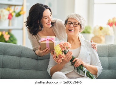 Chúc mừng ngày của mẹ! Người phụ nữ trẻ đẹp và mẹ cô ấy với hoa và hộp quà ở nhà.