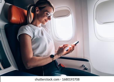 Turista en gafas clásicas que revisan el correo electrónico recibido en las redes sociales durante la mensajería de Internet a bordo en un avión de pasajeros, una mujer caucásica reserva boletos de regreso conectados a wifi de alta velocidad