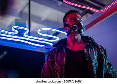 Jugador de cyberpunk de raza mixta en máscara de pie cerca de la iluminación de neón y mirando a otro lado