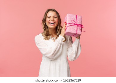 Concepto de vacaciones, celebración y mujeres. Retrato de una chica rubia carismática feliz sacudiendo una caja de regalo preguntándose qué hay dentro mientras celebra cumpleaños, recibe regalos de cumpleaños, fondo rosa