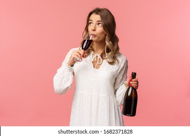 Concepto de vacaciones, primavera y fiesta. Retrato de mujer rubia linda e independiente despreocupada bebiendo vino de vidrio, sosteniendo una botella y degustando una bebida, mirando hacia arriba, celebrando con amigos, fondo rosa