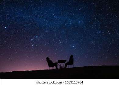pareja joven sentada en una mesa en una duna del desierto mientras habla, se relaja y observa las estrellas y la vía láctea sobre ellos