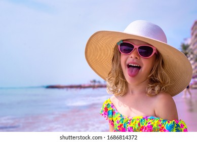Happy little girl makes a funny grimace and shows her tongue. Adorable girl in a big straw hat, sun glasses and in a swimsuit relaxing on the beach near sea. Cute child on vacation looks at the sea.