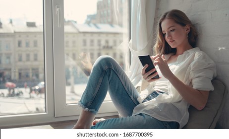 O femeie destul de tânără care stă pe pervazul ferestrei acasă și trimite mesaje pe telefonul ei.