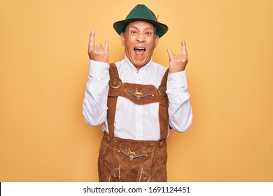 Senior grey-haired man wearing german traditional octoberfest suit over yellow background shouting with crazy expression doing rock symbol with hands up. Music star. Heavy music concept.
