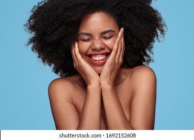Positive young black female with bare shoulders and beautiful skin touching face and laughing with eyes closed on blue background