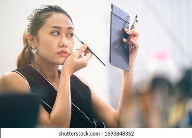 Retrato lateral de la actriz modelo asiática mirando el espejo y tratando de maquillarse con un enfoque selectivo, la industria de modelos conceptuales, el estilo de vida de la actriz modelo, el maquillaje natural, la industria del entretenimiento.