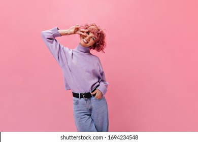 Schattige, vrolijke vrouw in paarse trui en spijkerbroek toont vredesteken. Charmante dame in spijkerbroek die lacht op geïsoleerde roze achtergrond.