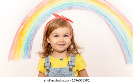 Nettes 2-jähriges Mädchen schaut in die Kamera. Mädchen in bunter Kleidung malt mit Kreide einen Regenbogen an die Wand. Kinderaktivitäten zu Hause. Zeichen der Hoffnung. Coronavirus.