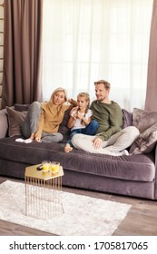 Captivated family of mother, father and little daughter watching movie together sitting on comfortable sofa at home