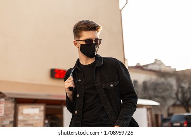 Pria muda hipster tampan dengan gaya rambut berkacamata hitam trendi dengan jaket denim hitam modis dengan topeng hitam medis bergaya berjalan di jalan pada hari yang cerah. Model pria yang menarik. Mode 2020. Musim semi.