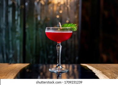 Een roodzure cocktail in een coupéglas, versierd met een dennentak op tafel in een bar