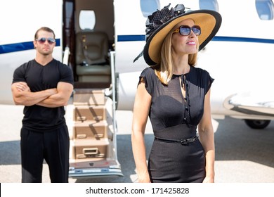 Glückliche Frau mit Sonnenhut und Sonnenbrille mit Leibwächter und Privatjet im Hintergrund