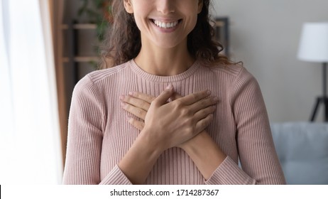 Close up focus on happy sincere female holding folded hands on chest. Emotional positive kind candid millennial woman feeling thankful indoors, showing gratitude sign, believe faith charity concept.