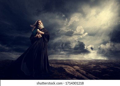 Monje sosteniendo la Biblia mirando hacia Dios Sky Light, viejo sacerdote con túnica negra en Storm Mountains