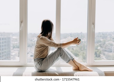 Young woman looking through the window with a city view, sitting on a windowsill, drinking coffee or tea in the morning.