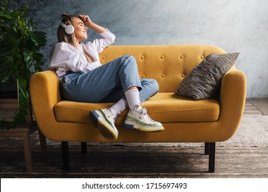 Hermosa mujer rubia con auriculares blancos se sienta en un sofá amarillo y disfruta escuchando música, podcast.