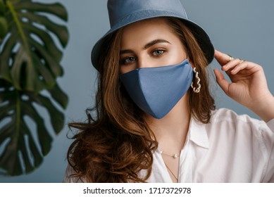 Woman wearing stylish outfit with luxury designer protective blue face mask, bucket hat, pearl earrings. Trendy Fashion accessory during quarantine of coronavirus pandemic. Close up studio portrait