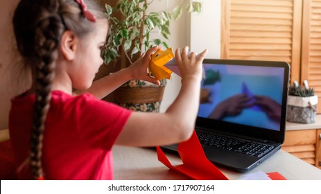 Cute little elementary schoolgirl doing origami fish with folded color paper looking video on laptop, online workshop, kids at-home activity, creativity and distant education, focus on hands