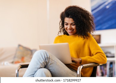 Afbeelding van een vrolijke Afro-Amerikaanse vrouw die een laptop gebruikt terwijl ze op een stoel in de woonkamer zit