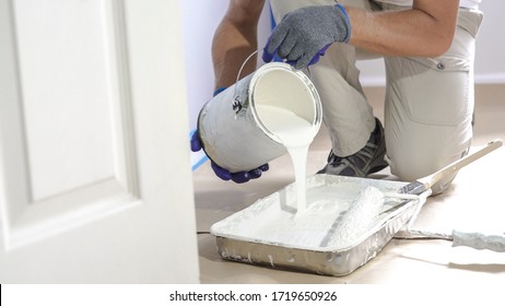 Seorang pria menuangkan cat ke dalam baki dan mencelupkan rol. Pekerja konstruksi interior profesional menuangkan cat warna putih ke baki.