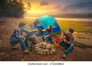 Fröhliche Freunde beim Camping, die Musik hören und ein Lagerfeuer in der Natur am See genießen. Ein Reisender campt in den Bergen und lässt den Sonnenuntergang bei einem Drink auf sich wirken. (Bildunterschriften: Rauschen und Filmkorn/Weichzeichner)