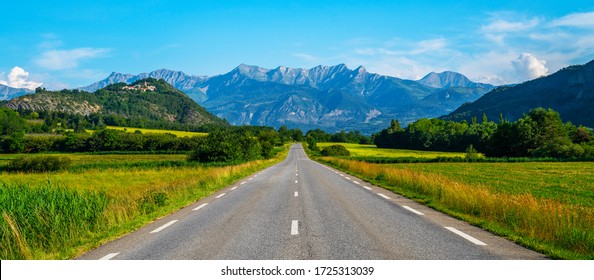 Jalan lurus panjang menuju pegunungan di Prancis. Pemandangan musim semi dan musim panas yang cerah dan penuh warna. Bidang kuning pemerkosaan berbunga dan langit biru dengan awan. Pemandangan alam, Eropa.