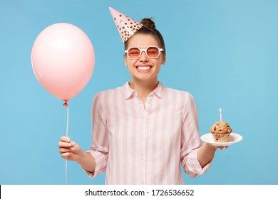 Smiling woman celebrating her birthday with pink balloon and cake in hands, wearing festive hat, enjoying surprise from friends, isolated on blue background