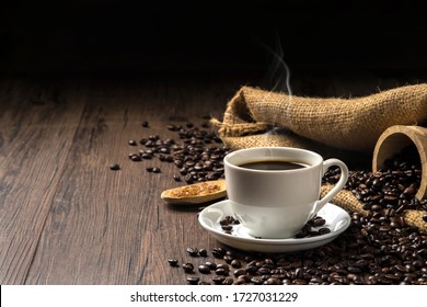 Heißer Kaffee in einer weißen Kaffeetasse und viele Kaffeebohnen und Zucker auf einem Holztisch in einer warmen, hellen Atmosphäre, auf dunklem Hintergrund, mit Kopierraum.