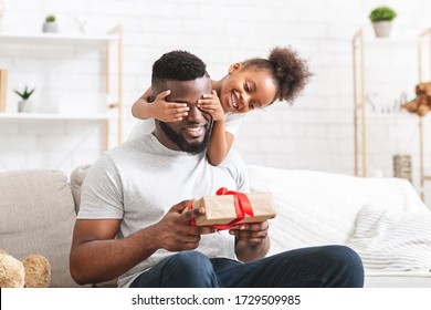 Süßes kleines Afro-Mädchen, das am Vatertag die Augen ihres Vaters bedeckt, Papa hält Geschenkbox, Inneneinrichtung, freier Raum