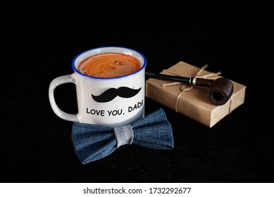 Mug of coffee with paper mustache, denim bow tie, gift box and smoking tobacco pipe on black background. Happy father's day concept. Inscription Love you dad. Greeting card