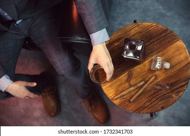 Caballero toma un vaso de whisky y una mesa de madera con jarra de whisky y puros. Vista desde arriba.