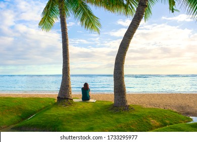 One teenage girl sitting alone on empty Hawaiian beach in Honolulu between two coconut trees looking out at ocean with back towards camera at sunset