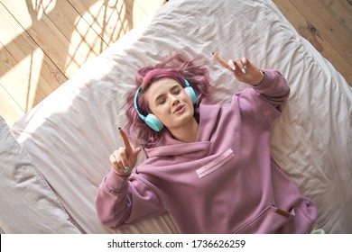 Happy funny teen girl with pink hair wear headphones lying in comfortable bed listening new pop music enjoying singing song with eyes closed relaxing in cozy bedroom at home. Top view from above.
