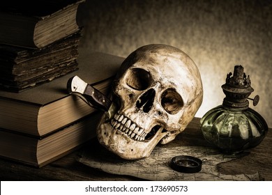 Still life, human skull with knife in the mouth beside old book, old compass on torn map and ancient lantern