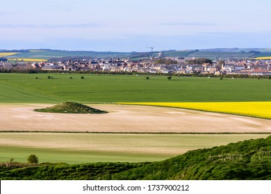 An ancient round barrow stands in a farm field in Dorset, with new build houses and construction of the new Dorchester suburb of Poundbury in the background.
