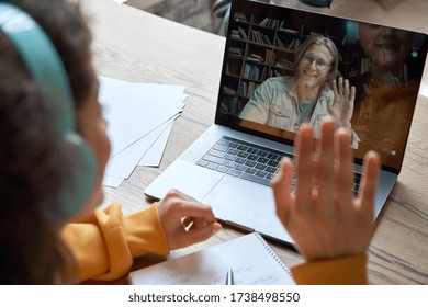 Испаноязычная девушка-подросток, студентка колледжа, обучающаяся дистанционно, машет рукой, занимаясь с онлайн-преподавателем на экране ноутбука. Видеозвонок по Zoom для электронного обучения, видеоконференция с репетитором. Вид через плечо.