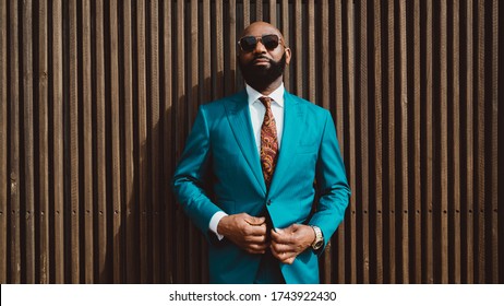 Un apuesto hombre africano maduro, calvo y barbudo, con gafas de sol y un elegante traje azul o verde azulado con corbata, está parado frente a una pared hecha de madera rayada y abrochando un botón de traje