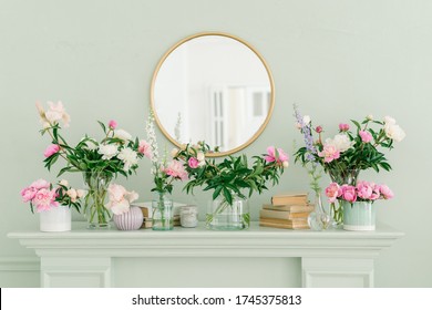 Roze en witte pioenrozen op de boekenplank in glazen vazen. Ronde gouden spiegel aan de muur boven de open haard