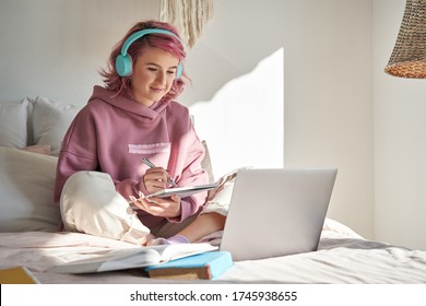 Hipster teen girl school student with pink hair wear headphone write notes watch video online webinar learn on laptop sit in bed distance elearning course video conference pc call in bedroom at home.