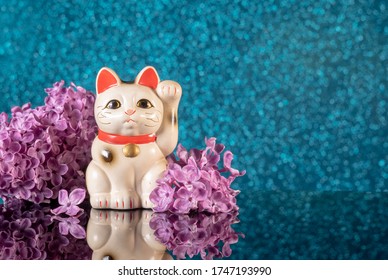 Japoński symbol szczęścia – kot Maneki Neko otoczony kwiatami bzu na niebieskim tle bokeh z odbiciem