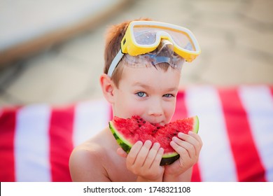 Little blue-eyed blond boy wearing yellow Diving Goggles eating watermelon sitting on the striped red and white beach towel in the summer